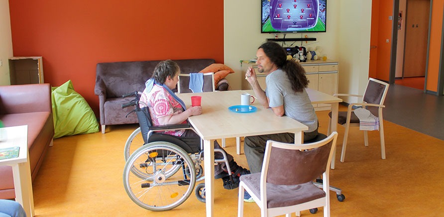 Eine Rollstuhlfahrerin und ein Mann sitzen sich gegenüber an einem Holztisch. Auf dem Tisch stehen ein Teller, ein Becher und eine Kaffeetasse. Die Personen befinden sich in einem Gemeinschaftsraum mit zwei Sofas. Im Hintergrund leuchtet ein Fernsehbildschirm.